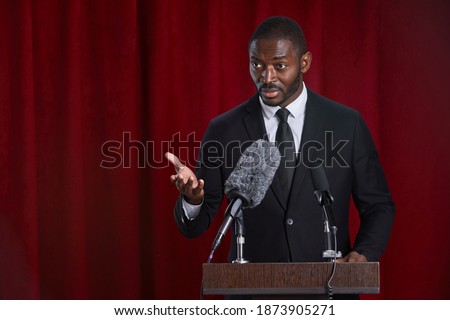 Waist up portrait of African-American man speaking to microphone standing at podium on stage against red curtain, copy space Royalty-Free Stock Photo #1873905271