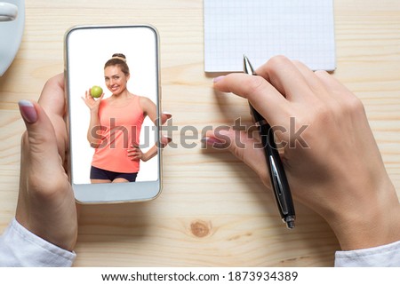 Fitness coaching and nutrition advice online. Female hands wirh smartphone, friendly smiling caucasian woman in sportswear with green apple on the screen, selective focus. New reality, distance