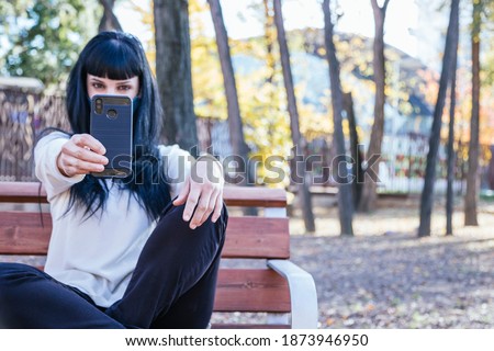 Brunette girl with mask holding her smartphone sitting on a park bench. Girl with black pants and white sweater with straight hair. Hoorizontal image with copispace. Technology concept.