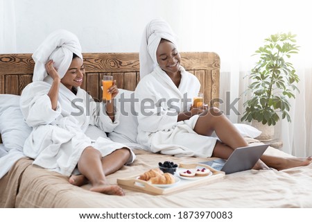 Two joyful young black women having breakfast in bed at hotel, watching romantic movie on laptop. African american pretty girlfriends in bathrobes having healthy snack in bedroom, free space