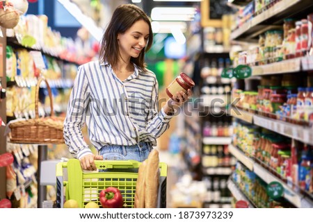 Portrait Of Smiling Woman With Shopping Cart In Supermarket Buying Groceries Food Walking Along The Aisle And Shelves In Grocery Store, Holding Glass Jar Of Sauce, Choosing Healthy Products In Mall Royalty-Free Stock Photo #1873973239