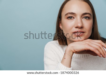 Close-up portrait of young smiling attractive woman posing isolated over blue background, holding hand at her chin