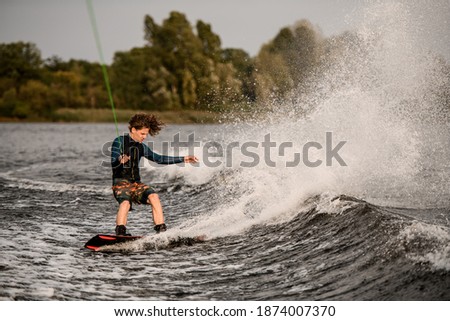 young athletic man holding taut rope with his hands and rides down on wave on wakeboard. Coastline with green trees in background.