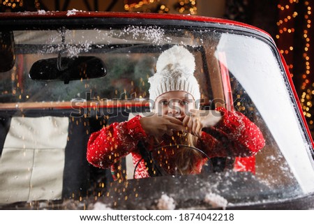 Blonde teenage girl in a white knitted hat with a pom-pom behind the glass in a red car. Selective focus. Blurry lights.