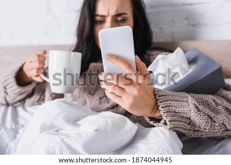sick young brunette woman with hot drink and tissue holding smartphone in bed, blurred background