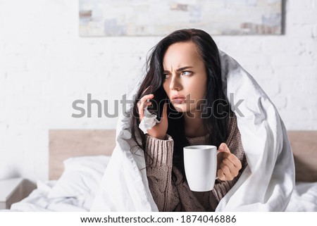 sick young brunette woman with hot drink and tissue talkign on smartphone in bed