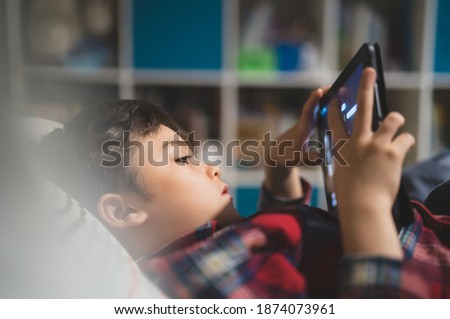Soft focus young kid playing game alone on tablet, Cute boy watching cartoons on digital pad while lying in bed, Candid shot Happy child relaxing and enjoying on his own in bed room in the morning. 
