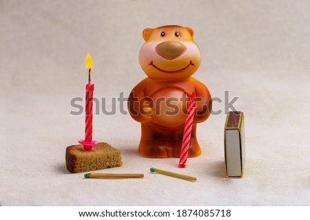 Toy bear next to a burning candle stuck in a piece of bread and matches on a linen cloth close up