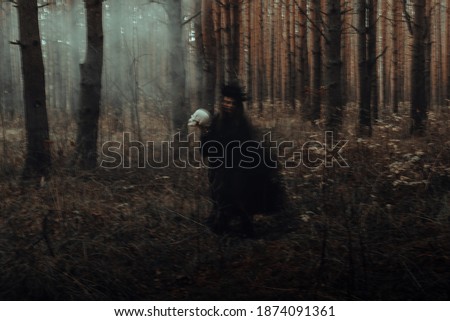 blurred silhouette of a terrible witch with a skull in her hands performing a mystical occult satanic ritual in a dark gloomy forest