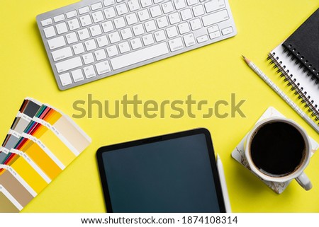 Designer desk top view with color guide palette, graphic tablet, coffee cup, paper notebook, keyboard on yellow background. Flat lay, view from above. Creative professional workspace.