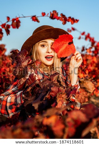 Beautiful young girl in a hat. Behind it is a background of red grape leaves. Autumn photo, a lot of emotions on her face, a grape leaf in her hand, she closes one eye with it and smiles beautifully.