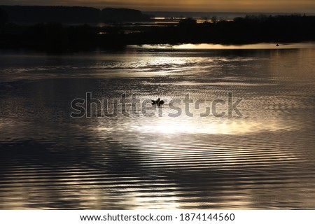Night river.The black sky with Golden stripes is reflected in the water with ripples on the surface.In the center of the water area is a distant silhouette of a fisherman in a boat in the moonlight