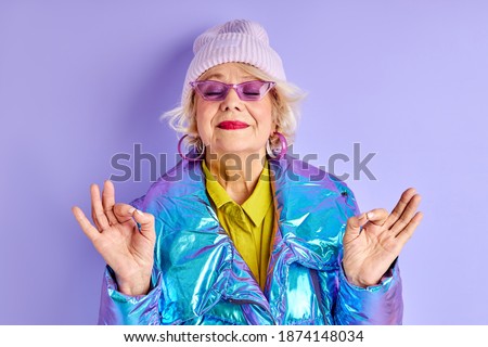 senior woman in stylish wear meditating isolated over purple background, attractive woman keeps calm, in yoga pose