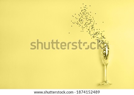 Star-shaped silver gray confetti poured out of champagne glass on illuminating yellow background with copy space for text. Holiday and celebration concept. Main color trend of the year of 2021.
