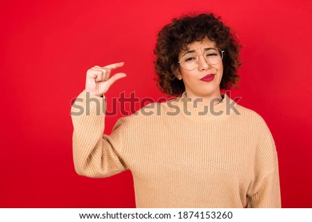 Upset Young beautiful Arab woman wearing knitted sweater standing against red background shapes little gesture with hand demonstrates something very tiny small size. Not very much