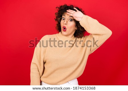 Young beautiful Arab woman wearing knitted sweater standing against red background wiping forehead with hand making phew gesture, expressing relief feels happy that he prevented huge disaster. 