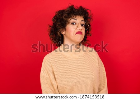 Young beautiful Arab woman wearing knitted sweater standing against red background with snobbish expression curving lips and raising eyebrows, looking with doubtful and skeptical expression, suspect a Royalty-Free Stock Photo #1874153518