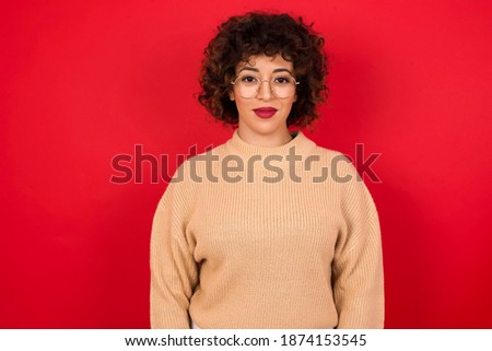 Happy Young beautiful Arab woman wearing knitted sweater standing against red background looking at camera with charming cute smile.