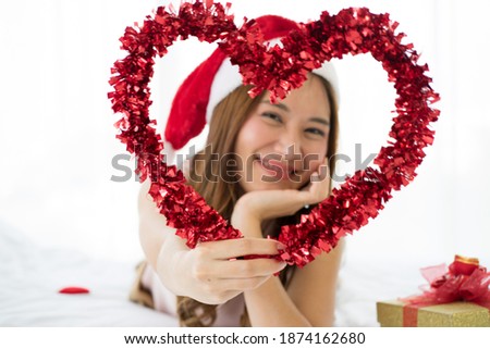 selected focus of red heart frame and blurred image of Asian Attractive  young Wolman on bed wearing red Santa hat and red gift box during Christmas holiday. new normal concept. 