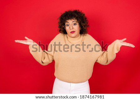 Puzzled and clueless Young beautiful Arab woman wearing knitted sweater standing against red background with arms out, shrugging shoulders, saying: who cares, so what, I don't know. Negative human emo