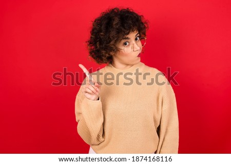 No sign gesture. Closeup portrait unhappy Young beautiful Arab woman wearing knitted sweater standing against red background raising fore finger up saying no. Negative emotions facial expressions.