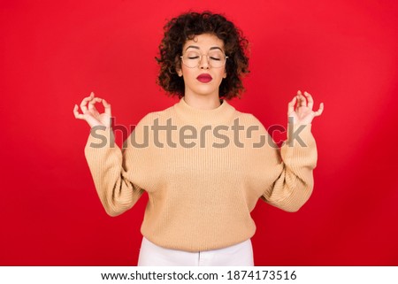 Young beautiful Arab woman wearing knitted sweater standing against red background doing yoga, keeping eyes closed, holding fingers in mudra gesture. Meditation, religion and spiritual practices.