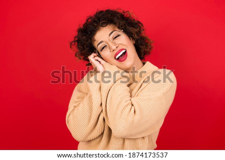 Dreamy Young beautiful Arab woman wearing knitted sweater standing against red background with pleasant expression, closes eyes, keeps hands crossed near face, thinks about something pleasant