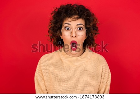 Young beautiful Arab woman wearing knitted sweater standing against red background having stunned and shocked look, with mouth open and jaw dropped exclaiming: Wow, I can't believe this. Surprise and 