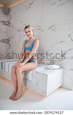 Full length portrait of happy cheerful blonde woman in bikini sitting on the white granite bench in spa resort
