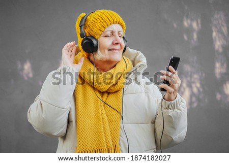 Elderly female listening to music against violet  wall. Senior woman in stylish outerwear listening to music in headphones and dancing against violet wall on city street