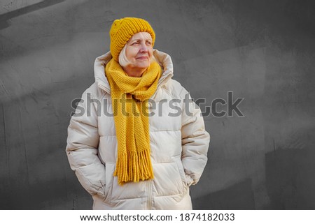 Stylish aged woman in warm coat with yellow knitted scarf and hat   looking away while standing against light blue  wall on sunny day