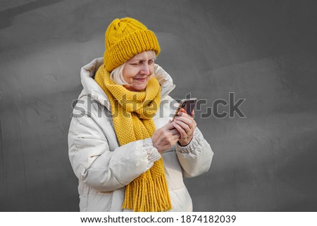 Modern senior woman in outerwear using smartphone. Contemporary elderly female in trendy outerwear smiling and browsing mobile phone while standing against blue wall on street