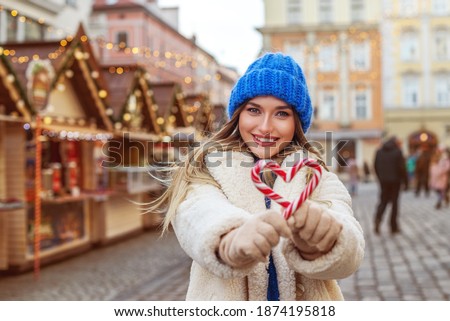 Christmas, New Year, winter holidays conception: happy smiling lady posing at street festive fair, holding candy canes. Funny girl makes heart with lollipops. Copy, empty space for text
