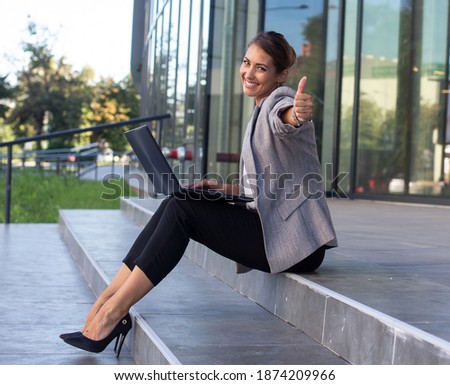 Pretty young business woman sitting on stair in front of office building, holding laptop in lap, having conference call and showing thumb up