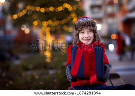 smiling boy holding christmas present enjoying outdoor holiday time with beautiful decorated with lights tree in the background, copy space on left, family holiday concept