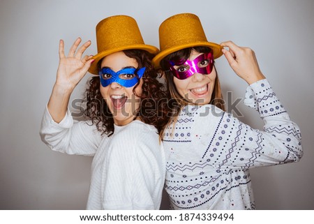 
Two beautiful women saying goodbye to the year 2020 in pajamas from home due to the pandemic caused by covid19. Festive and fun attitude wearing golden hat and colorful mask