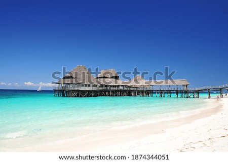 A building on pillars, against a background of blue sky and blue, transparent ocean.