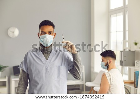 Young black man doctor nurse in medical face fask holding syringe with vaccination against COVID-19 and waiting man patient at background in medical clinic. Vaccine, epidemic, virus protection concept