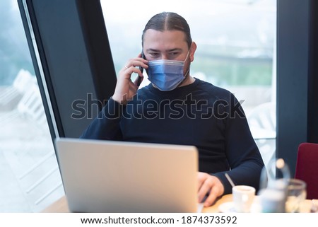 Businessman in medical mask during quarantine. A young man in a cafe works at a laptop by the window. Isolation, coronavirus, work at the home.
