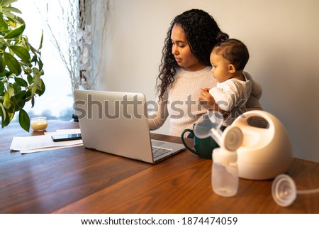 A beautiful young mixed Race African American mother holds her daughter while taking notes at her dining table serving as a temporary remote work from home station with breast pump in foreground. Royalty-Free Stock Photo #1874474059