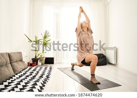 Young calm woman wearing homewear practicing yoga in warrior pose at the light living room. Stock photo. Virabhadrasana concept