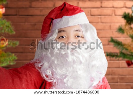 Asian man in Santa costume taking a selfie with brick wall background. Merry Christmas