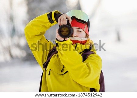 Female photographer in ski equipment takes pictures with a camera with a huge lens. Winter clothes: red pants and a warm yellow jacket, knitted hat, ski goggles. Selective focus.