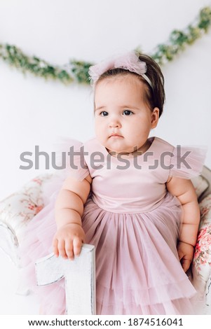 a little cute girl in a beautiful dress is sitting in a chair, holding the number 1 in her hand. first birthday, children's day