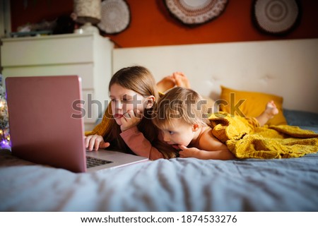 Girls children in bedroom are looking at screen near Christmas tree in real interior, concept of Christmas sales and interesting cartoons. siblings on the bed with blanket lying looking laptop