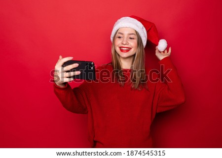 Surprised young Santa woman in Christmas hat doing selfie shot on mobile phone isolated on red background.