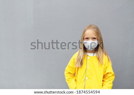 little caucasian girl 7 years old with blonde hair in yellow raincoat and face mask during Coronavirus pandemic. Colors of the year 2021 ultimate gray and illuminating Royalty-Free Stock Photo #1874554594