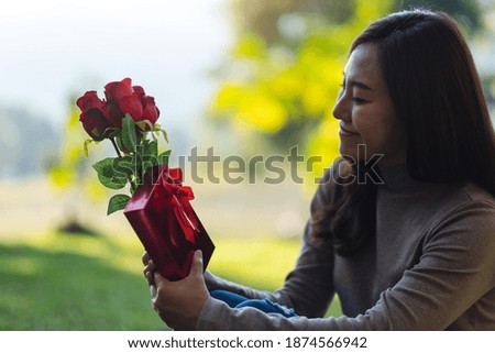 Closeup image of a beautiful asian woman holding red roses flower and a gift box in the park