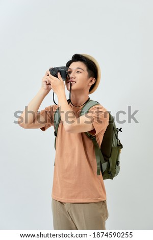 Happy handsome young Asian man taking photo while wearing hat and backpack over white background.