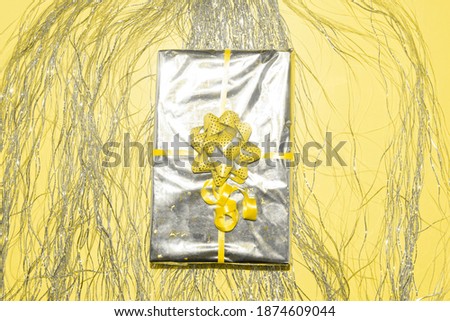 Christmas present wrapped in silver holographich foil with yellow ribbon bow on tinsel fringe and illuminating background